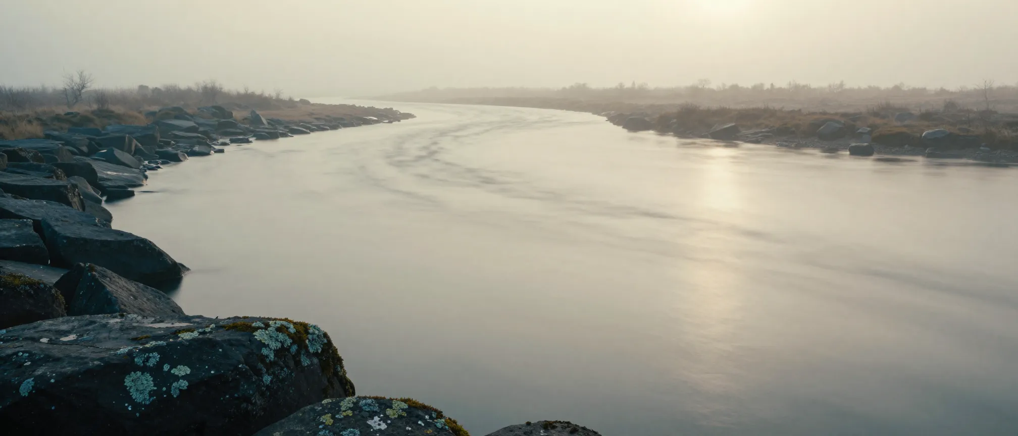 A wide river bending through mist at dawn, lichen-flecked stones along the near bank.